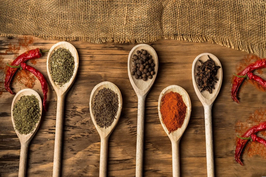 Wooden spoons with spices like chili, cumin, and herbs, set on a rustic wooden surface.