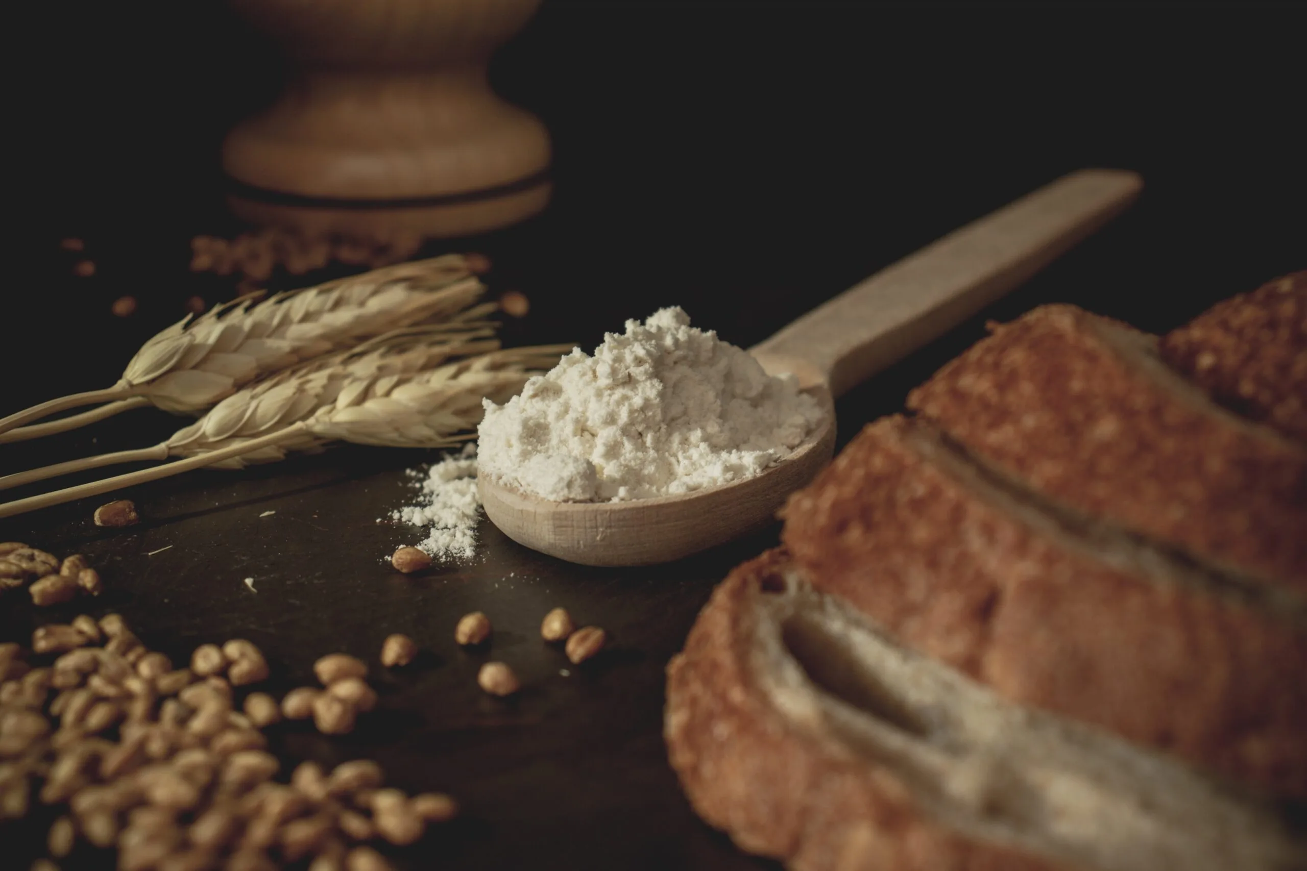 Rustic bread slices, wheat stalks, and a wooden spoon with flour on a dark surface.